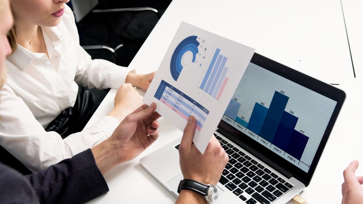 Two colleagues at a white desk reviewing printed blue bar charts beside an open laptop