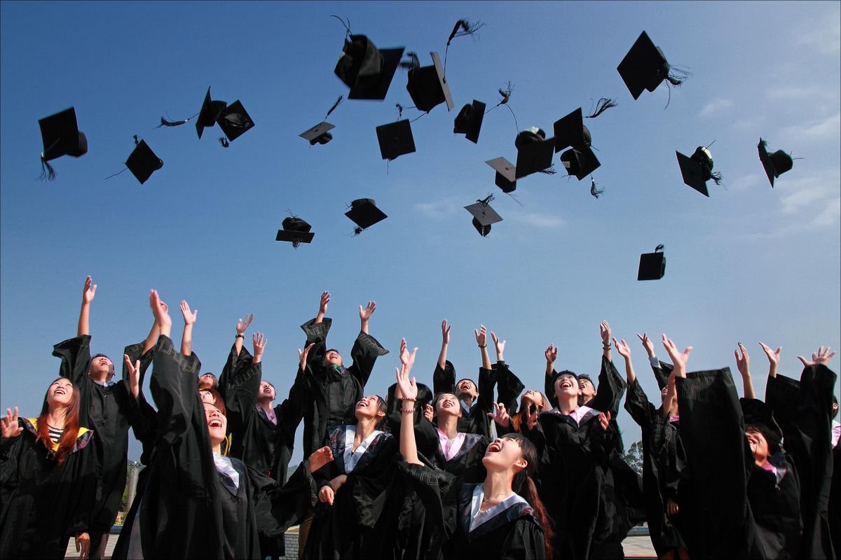 Group of graduates in black gowns tossing mortarboard caps into a clear blue sky